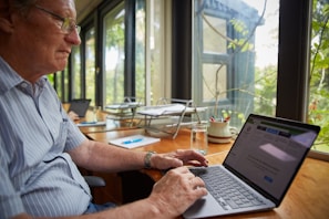 An elderly person wearing a striped shirt is working on a laptop at a wooden desk by a large window. The desk also contains papers, a glass of water, and a mug filled with pens. The view outside the window displays a mix of greenery.