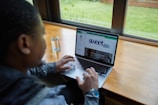 A person is using a laptop while seated at a wooden table beside a window. Outside the window, there is a view of a green lawn. The laptop screen displays a webpage with the title 'sweetlife'. A glass of water is placed on the table next to the person.