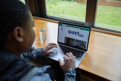 A person is using a laptop while seated at a wooden table beside a window. Outside the window, there is a view of a green lawn. The laptop screen displays a webpage with the title 'sweetlife'. A glass of water is placed on the table next to the person.