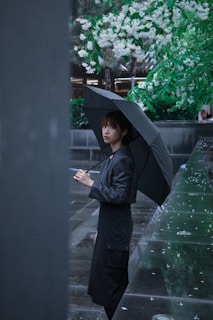 A person stands under a black umbrella, wearing a dark raincoat, on a rainy day. There are green trees with white blossoms overhead and the ground is wet from the rain.