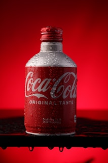 A chilled bottle of Coca-Cola is presented against a bright red background. The bottle is adorned with droplets of condensation, highlighting its cold and refreshing nature. The iconic red and white branding is prominently displayed on the label.