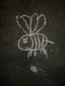 A simple chalk drawing of a bee on a dark, rough asphalt surface. The bee has three visible wings, a large eye, and stripes on its body.