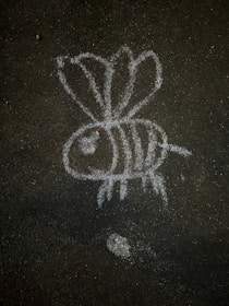 A simple chalk drawing of a bee on a dark, rough asphalt surface. The bee has three visible wings, a large eye, and stripes on its body.