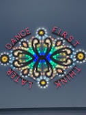 A decorative neon sign featuring illuminated text and intricate patterns with vibrant colors. The words 'DANCE FIRST THINK LATER' are arranged around a symmetrical design that includes curved lines and circular elements.