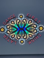 A decorative neon sign featuring illuminated text and intricate patterns with vibrant colors. The words 'DANCE FIRST THINK LATER' are arranged around a symmetrical design that includes curved lines and circular elements.