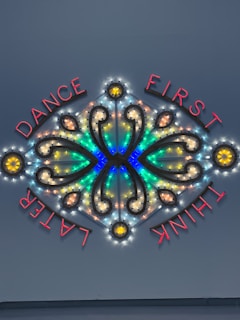 A decorative neon sign featuring illuminated text and intricate patterns with vibrant colors. The words 'DANCE FIRST THINK LATER' are arranged around a symmetrical design that includes curved lines and circular elements.