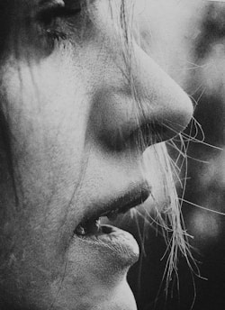 A close-up black and white photograph captures a person's face in profile, highlighting the texture of the skin and the intricacies of the lips. Fine strands of hair partially obscure the face, adding a sense of intimacy and softness to the image.