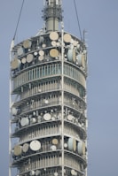 A close-up view of a tall telecommunications tower equipped with numerous satellite dishes and antennas arranged in circular tiers. The structure is metallic and industrial in design, showing intricate technical components.