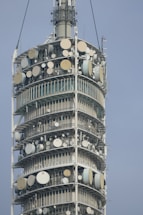 A close-up view of a tall telecommunications tower equipped with numerous satellite dishes and antennas arranged in circular tiers. The structure is metallic and industrial in design, showing intricate technical components.