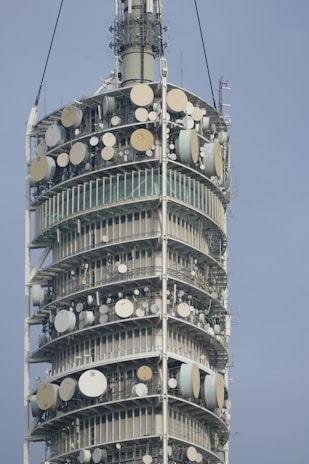 A close-up view of a tall telecommunications tower equipped with numerous satellite dishes and antennas arranged in circular tiers. The structure is metallic and industrial in design, showing intricate technical components.