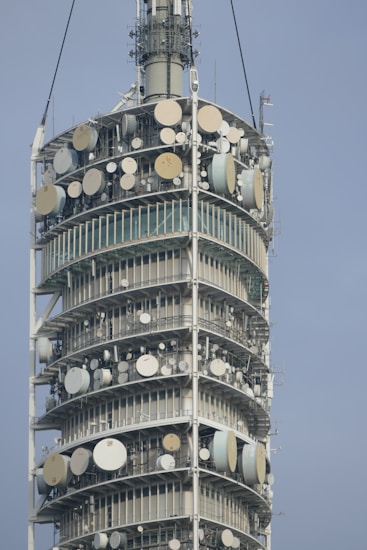 A close-up view of a tall telecommunications tower equipped with numerous satellite dishes and antennas arranged in circular tiers. The structure is metallic and industrial in design, showing intricate technical components.