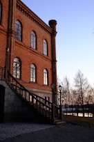 A historic brick building with tall arched windows and a decorative chimney is seen at sunset. A stone staircase with a wrought iron railing ascends along the side of the building. There are vintage street lamps along the path, and bare trees are silhouetted against the blue and pink sky.