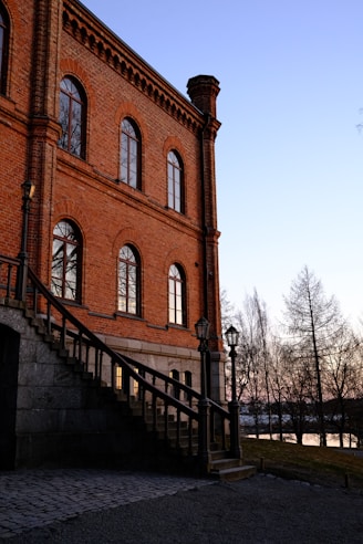 A historic brick building with tall arched windows and a decorative chimney is seen at sunset. A stone staircase with a wrought iron railing ascends along the side of the building. There are vintage street lamps along the path, and bare trees are silhouetted against the blue and pink sky.