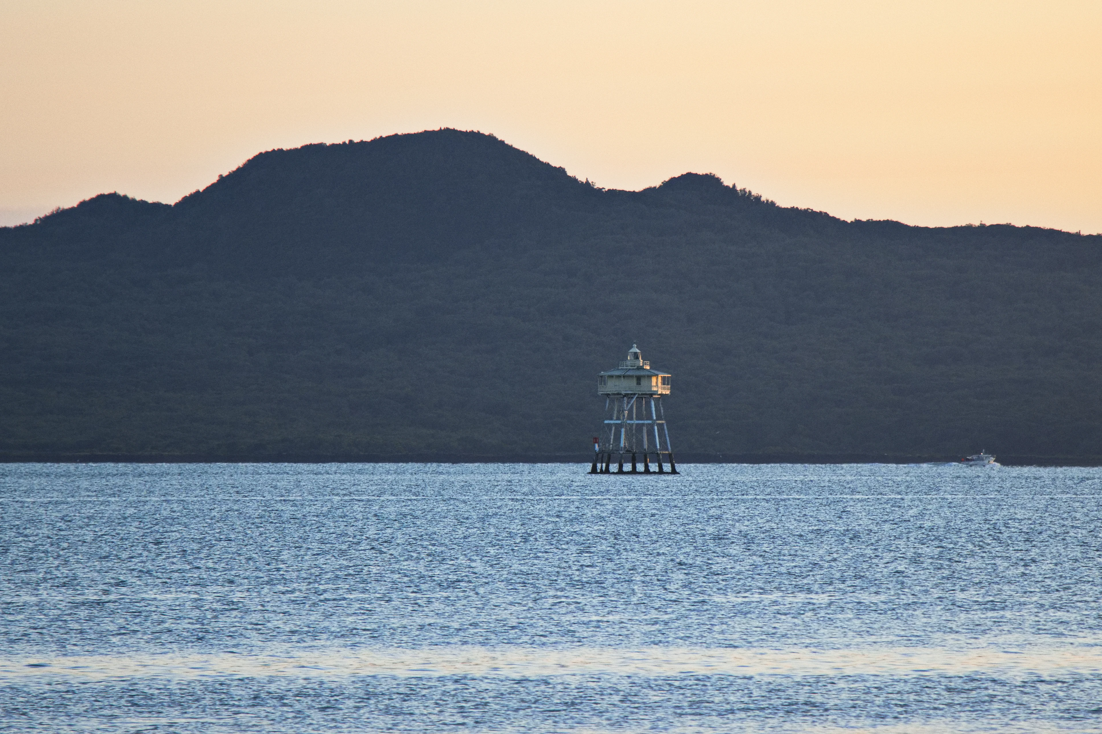 A navigation beacon stands in calm harbour water at sunset, silhouetted hills behind it.