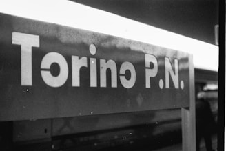 A monochromatic photograph of a sign reading 'Torino P.N.' with a blurred background and part of a person visible on the right.