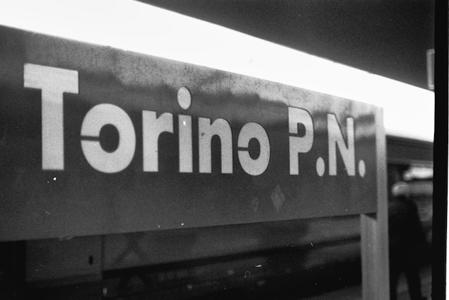 A monochromatic photograph of a sign reading 'Torino P.N.' with a blurred background and part of a person visible on the right.