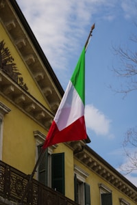 An Italian flag with green, white, and red vertical stripes is affixed to the side of a yellow building with dark green shutters. The building features classic architectural details such as a decorative metal railing and intricate cornices. The scene includes a partly cloudy sky and some bare tree branches.
