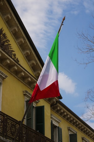 An Italian flag with green, white, and red vertical stripes is affixed to the side of a yellow building with dark green shutters. The building features classic architectural details such as a decorative metal railing and intricate cornices. The scene includes a partly cloudy sky and some bare tree branches.