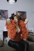 A person with bold makeup and red hair is wearing a vibrant orange feathered shawl over a deep red bodysuit and black gloves. The setting appears to be a contemporary living room with abstract wall art and modern furniture.
