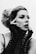 A black and white portrait of a person with styled hair and dramatic makeup, including winged eyeliner and bold lipstick. The individual wears long, lace, fingerless gloves and poses with hands framing their face, exuding a sense of elegance and contemplation.