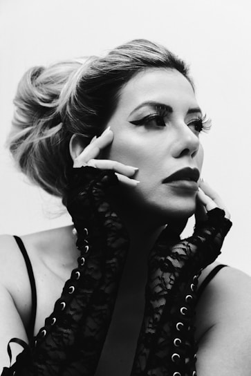 A black and white portrait of a person with styled hair and dramatic makeup, including winged eyeliner and bold lipstick. The individual wears long, lace, fingerless gloves and poses with hands framing their face, exuding a sense of elegance and contemplation.
