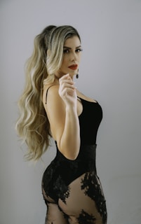 A woman with long, wavy blonde hair is wearing a black, form-fitting outfit featuring lace details and a sheer fabric. She is posing confidently against a neutral background, looking over her shoulder.
