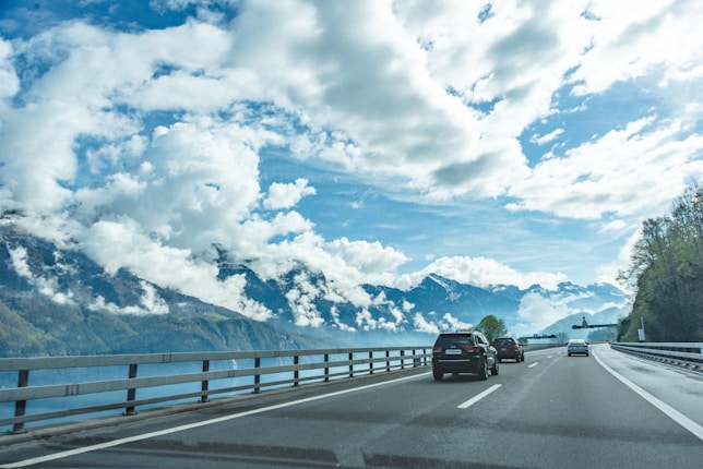 A scenic highway stretches ahead, flanked by guardrails, with several cars traveling along it. To the left, a large body of water lies serenely beneath a range of majestic, snow-capped mountains enveloped in fluffy white clouds. The sky above is a bright blue with scattered clouds, creating a picturesque and serene atmosphere. Lush trees line the right side of the road.
