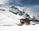 A wooden cabin sits near the base of snow-covered mountains with pathways and ski tracks visible on the white slopes. The sky is cloudy with patches of blue, and a few people can be seen walking in the snow.