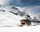 A wooden cabin sits near the base of snow-covered mountains with pathways and ski tracks visible on the white slopes. The sky is cloudy with patches of blue, and a few people can be seen walking in the snow.