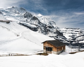A wooden cabin sits near the base of snow-covered mountains with pathways and ski tracks visible on the white slopes. The sky is cloudy with patches of blue, and a few people can be seen walking in the snow.