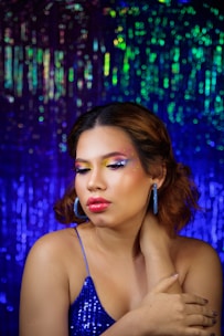 A woman with vibrant makeup featuring bright eyeshadow and glossy red lips is posing against a shimmering multicolored background. Her eyes are closed, and she wears a sparkly blue top and matching earrings. The scene is colorful and glamorous.