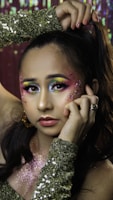 A person with elaborate and colorful makeup featuring yellow, blue, and pink hues blended around the eyes. Glitter accents are applied on the cheeks, and the individual is wearing a sequined green garment. Their hair is styled up, with a hand holding it. The background is sparkling, complementing the festive look.