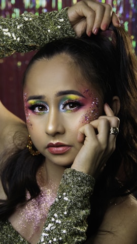 A person with elaborate and colorful makeup featuring yellow, blue, and pink hues blended around the eyes. Glitter accents are applied on the cheeks, and the individual is wearing a sequined green garment. Their hair is styled up, with a hand holding it. The background is sparkling, complementing the festive look.