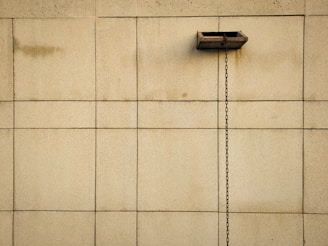 A beige tiled wall with a central horizontal duct from which a metal chain hangs vertically. The tiles are square and uniformly spaced, providing a symmetric and minimalistic appearance.