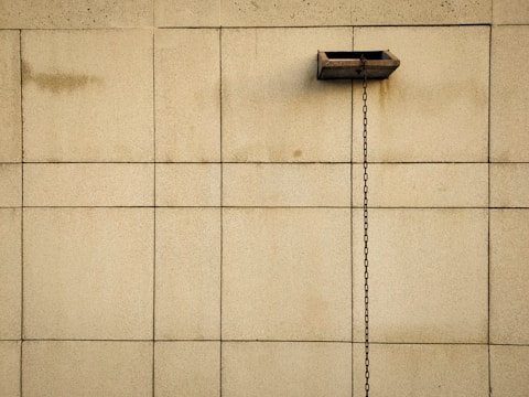 A beige tiled wall with a central horizontal duct from which a metal chain hangs vertically. The tiles are square and uniformly spaced, providing a symmetric and minimalistic appearance.