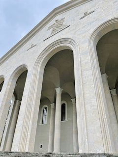 A view of a large, classical architectural structure featuring imposing arches and columns. The facade is embellished with stone carvings and statues, giving a sense of grandeur. The structure's surface has a light, beige hue and there are tall rectangular windows, adding to the building's intricate detailing.