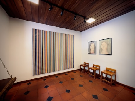The room features a wooden ceiling with track lighting. On the walls, there is a large painting with vertical stripes in various colors. Two framed portraits of faces are also displayed. Below them are three simple wooden chairs. The floor is tiled with a pattern of terracotta and black tiles.