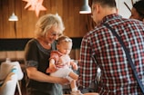 An elderly woman with glasses holds a young child in her arms, smiling at the child who is wearing a headband. A man in a plaid shirt is turned away from the camera, engaging with the woman and child. The setting is indoors, with warm lighting from hanging lamps and wooden walls.