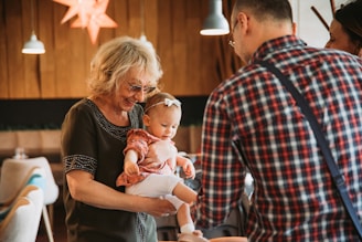 An elderly woman with glasses holds a young child in her arms, smiling at the child who is wearing a headband. A man in a plaid shirt is turned away from the camera, engaging with the woman and child. The setting is indoors, with warm lighting from hanging lamps and wooden walls.
