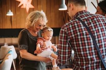 An elderly woman with glasses holds a young child in her arms, smiling at the child who is wearing a headband. A man in a plaid shirt is turned away from the camera, engaging with the woman and child. The setting is indoors, with warm lighting from hanging lamps and wooden walls.