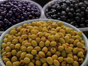 A close-up view of three bowls containing different types of olives. The foreground has a bowl filled with green olives, many of which are stuffed with red pimento. In the background, there are two bowls, one with dark purple olives and the other with black olives.