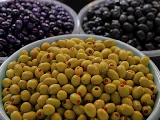 A close-up view of three bowls containing different types of olives. The foreground has a bowl filled with green olives, many of which are stuffed with red pimento. In the background, there are two bowls, one with dark purple olives and the other with black olives.