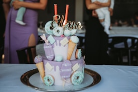 A two-tiered cake is adorned with light purple icing and various decorations including donuts, ice cream cones, lollipops, and cookies. The top of the cake has a golden name decoration that says 'Elena' and two sparkling candles. The cake is set on a silver tray on a white tablecloth. In the background, there are people, one holding a baby dressed in white and another wearing a purple dress, partially visible.