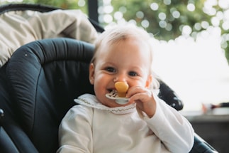 A baby with light hair is sitting in a dark-colored stroller or car seat. The baby is wearing a white outfit with a ruffled collar and is holding a pacifier or teething toy near their mouth. Blurred greenery is visible in the background, suggesting the setting might be outdoors.
