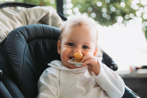 A baby with light hair is sitting in a dark-colored stroller or car seat. The baby is wearing a white outfit with a ruffled collar and is holding a pacifier or teething toy near their mouth. Blurred greenery is visible in the background, suggesting the setting might be outdoors.