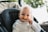 A baby with light hair is sitting in a dark-colored stroller or car seat. The baby is wearing a white outfit with a ruffled collar and is holding a pacifier or teething toy near their mouth. Blurred greenery is visible in the background, suggesting the setting might be outdoors.