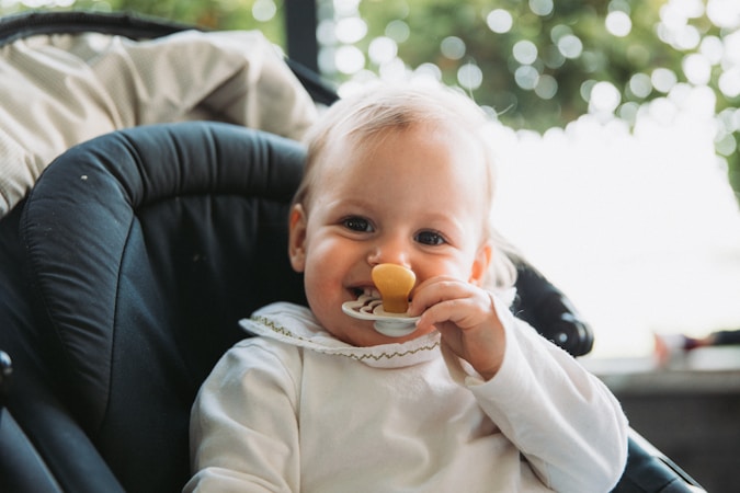A baby with light hair is sitting in a dark-colored stroller or car seat. The baby is wearing a white outfit with a ruffled collar and is holding a pacifier or teething toy near their mouth. Blurred greenery is visible in the background, suggesting the setting might be outdoors.