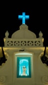 A religious structure with a lit blue cross at the top against a dark sky. Below the cross is a small alcove containing a statue of a saint, illuminated in a turquoise light. The facade of the building is ornate with decorative elements and has a classic architectural style.