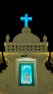A religious structure with a lit blue cross at the top against a dark sky. Below the cross is a small alcove containing a statue of a saint, illuminated in a turquoise light. The facade of the building is ornate with decorative elements and has a classic architectural style.