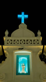 A religious structure with a lit blue cross at the top against a dark sky. Below the cross is a small alcove containing a statue of a saint, illuminated in a turquoise light. The facade of the building is ornate with decorative elements and has a classic architectural style.